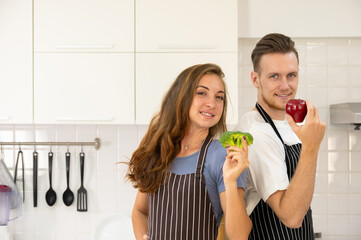 Portrait of smiling loving couple family standing at the kitchen. Male holding apple while female holding broccoli and looking at camera - 781138943