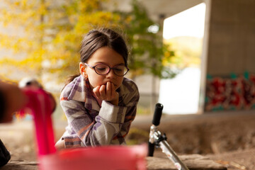 A tired little girl in a plaid shirt and glasses leaned her head on her hand while sitting under a bridge