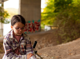Cute little girl in a plaid shirt and glasses sits sadly under a bridge
