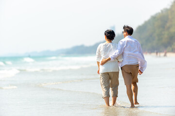 Happy Asian family senior couple walking together on tropical island beach at summer sunset. Retired elderly people relaxing and enjoy outdoor lifestyle travel nature ocean on holiday vacation.