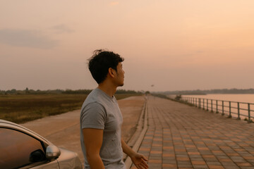 Asian man standing relaxing and leaning the car look at nature at sunset time, looking out the river, happy travel concept.