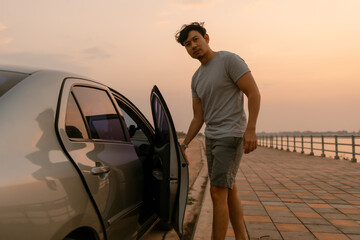 Asian man closing car door, parking a car next to the river at sunset time after long drive, happy life concept.