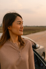 Vertical Close up of asian Thai woman standing next to the car and smiling look at the view near the river, sunset time, chilling traveling on vocation. 