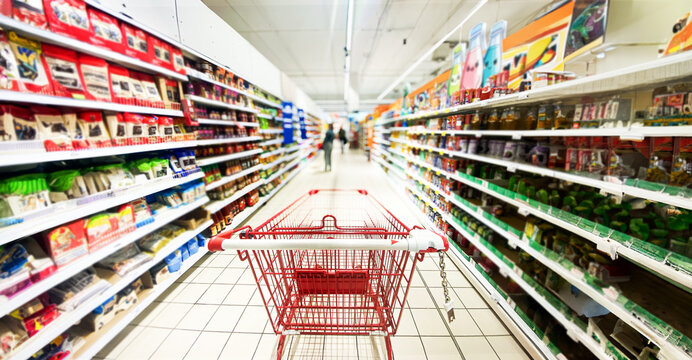 Pusty koszyk, w&oacute;zek sklepowy w supermarkecie, rozmazane tło, wnętrze sklepu, supermarketu, w&oacute;zek na zakupy, p&oacute;łki sklepowe z produktami. Hipermarket. Baner, miejsce na tekst.