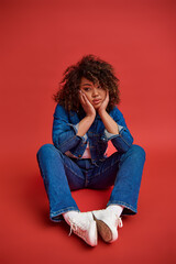 beautiful young african american woman in stylish denim outfit looking away on red backdrop