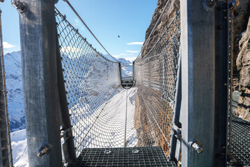 Obraz premium Metal suspension bridge with mesh flooring and cable support in mountainous terrain of Murren, Switzerland. Snowy landscape and Swiss Alps in the background.