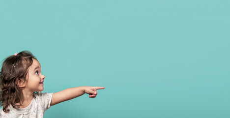 A little girl on a turquoise background points her finger to the side. Free space for text.
