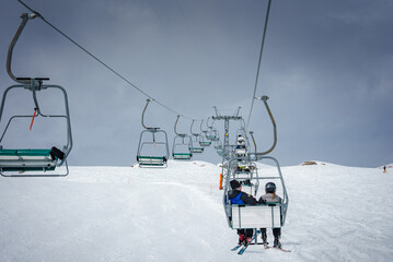 Ski lift taking skiers up snow covered mountain slope with overcast sky. Riders wear helmets,...