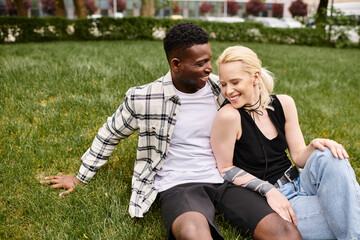 A happy, multicultural couple, consisting of an African American man and a Caucasian woman, sitting on the grass in a park.