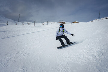 Snowboarder in action at snowy slope, dressed in winter sports gear with ski lift system in the background at a developed resort on a cold, overcast day.