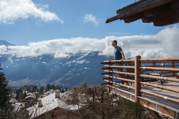 Man in casual attire standing on wooden balcony overlooking snow covered mountain village in the European Alps. Majestic snowy peaks in the background. - obrazy, fototapety, plakaty
