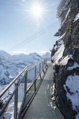 Metal walkway on rocky cliff at Murren ski resort, Switzerland. Snow covered mountains in distance, clear blue sky, and safe pathway for exploring Alps.