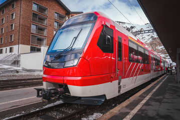 Modern red train at platform, likely part of Glacier Express in Switzerland. Sleek design with large front window for wide view. Snowy mountainous backdrop near Zermatt.