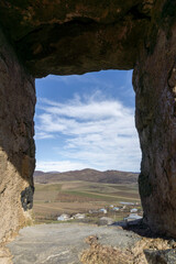 View of the village and mountains through the window from the Medieval fortress on a rock. Stone wall and agricultural fields.  Bright blue sky with clouds.  Kveshi fortress. Georgia