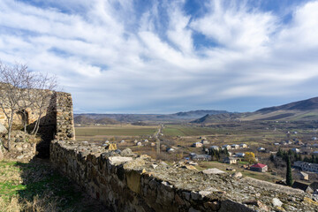 Fototapeta na wymiar View of the village and mountains from the Medieval fortress on a rock. Stone wall and part of tower. Bright blue sky with clouds. Kveshi fortress. Georgia