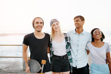Group, friends and portrait with hug on promenade by ocean for diversity, bonding or care at sunset. Men, women or gen z people with smile, embrace or relax on boardwalk with streetwear on vacation