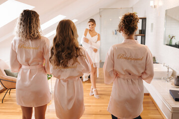 Three bridesmaids are dressed in robes with the inscription bridesmaid. The girls look at the bride standing with her back to the photographer.