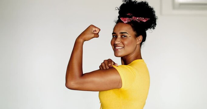 Happy Confident young African American woman showing strength, flexing arm looking at camera. One black latina shows muscle, we can do it symbol, wearing yellow shirt