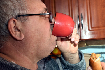 The picture shows an elderly man drinking from a cup and holding a pie in his other hand. Close-up.