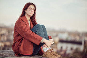 Pensive teenage girl in glasses with long red hair in red coat sits with his knees bent and looks away, against the background of sky and blurred cityscape.