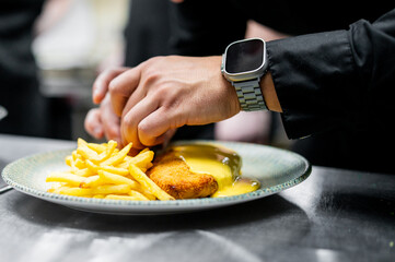 Close-up of a person in a professional kitchen garnishing a plate of crispy chicken and fries, wearing a smartwatch. Background suggests a professional kitchen setting