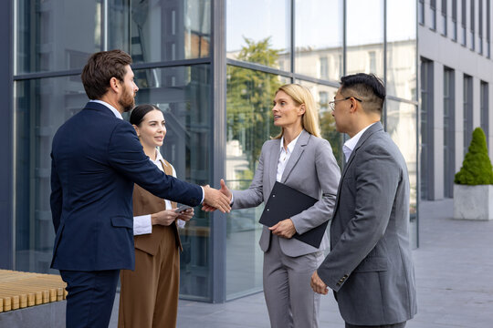 Interracial group of young business people standing on street outside office building in suits, man and woman shaking hands, making deal and getting to know partners and clients.