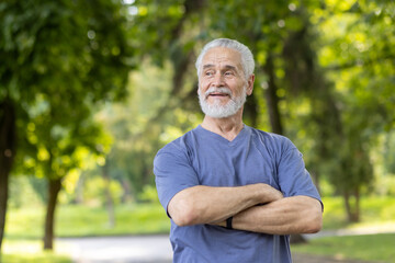 Naklejka na ściany i meble A close-up photo of an older gray-haired man standing in casual clothes in nature, crossing his arms on the ground and looking to the side with a smile.
