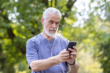Photo of an older gray-haired athletic man standing in the park wearing headphones and using a mobile phone.