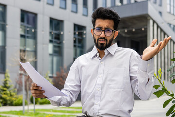 Portrait of an upset and disappointed young Indian man sitting outside a counter building, holding a document and waving his arms at the camera.