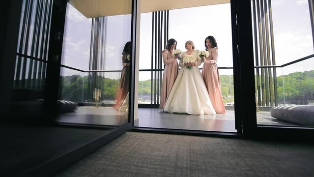 Bride with Bridesmaids Posing by Window, A smiling bride and bridesmaids in elegant dresses by a large window.