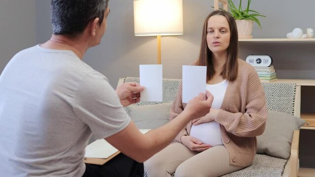 Professional therapy for mental health. Consultation with psychologist for counseling. Caucasian pregnant woman talking with therapist while sitting on sofa man showing pictures for patient