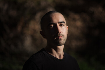 Chiaroscuro lighting of a young man with a buzz cut, looking at the camera with a neutral expression. He is posing against a completely defocused forest background.