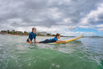 Happy boy learns to surf - young surfer learn to ride on surfboard with surf instructor on sea...