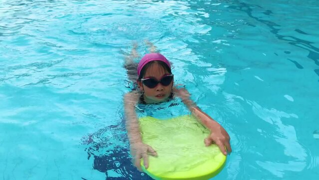Asian child girl using kick board in swimming pool, swimming direct to camera.