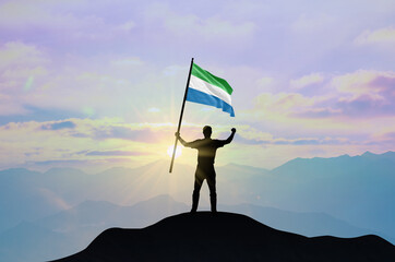 Sierra Leone flag being waved by a man celebrating success at the top of a mountain against sunset or sunrise. Sierra Leone flag for Independence Day.