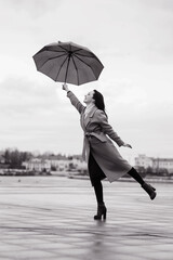 A girl in a light brown coat and a light scarf under an umbrella in black and white on a cloudy April evening against the background of Cheboksary