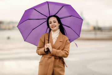 A girl in a light brown coat and a light scarf under a purple umbrella shot on a cloudy April evening against the background of Cheboksary