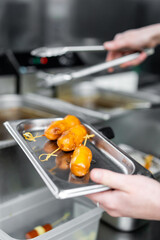 Professional chef preparing glazed appetizers in a kitchen. Golden brown, glossy appetizers are in focus on a stainless steel tray, with blurred kitchen equipment in the background