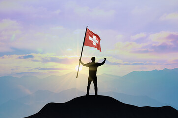 Switzerland flag being waved by a man celebrating success at the top of a mountain against sunset...
