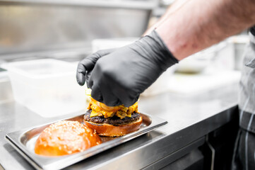 Close-up of a chef’s gloved hand assembling a juicy burger with various toppings on a metal tray in a professional kitchen setting