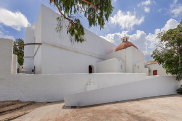 The exterior of Puig de Missa church in Santa Eulalia, Ibiza, captures the essence of Mediterranean architecture with its stark white walls and distinctive terracotta dome amidst lush greenery - obrazy, fototapety, plakaty