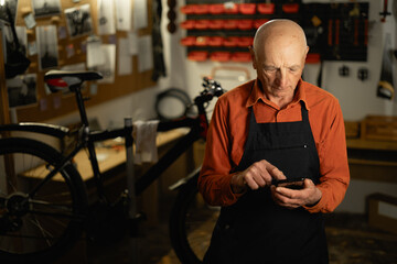 Senior Mechanic wearing apron in a bicycle repair shop using his mobile phone reading a text...