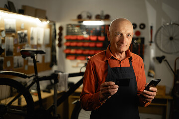 Old man repairing his bicycle standing holding credit card and mobile phone smiling at the camera