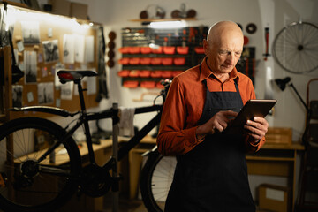 Old repairman using Technology on workplace of bicycle repair. Shot of man using a digital tablet...