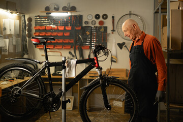 Mature male repairman repairing bicycle in garage or workshop, looking on wheel. Bike service, repair and upgrade - obrazy, fototapety, plakaty