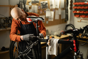 Old male mechanic working in bicycle repair shop, mechanic repairing bike. - obrazy, fototapety, plakaty