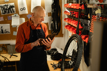 Old repairman uses a digital tablet while fixing a bike in a workshop while standing near a wheel. Bicycles repair and upgrade