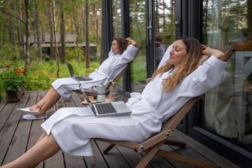 A women in a white bathrobe relaxes on a deck chair in a serene forest setting, working on a laptop with a cup of tea nearby, embodying the work-life balance