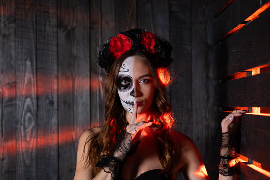 An enigmatic woman with Dia de los Muertos face paint poses mysteriously, red lighting enhancing the gothic atmosphere of the scene. The woman shows a gesture with her index finger covering her mouth