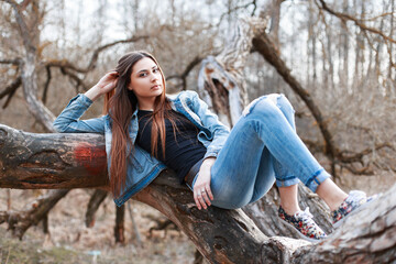 Beautiful fashion girl teenager model in a stylish denim outfit with floral sneakers lies on a tree...
